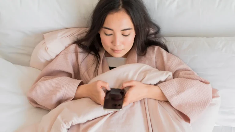 Woman in a pink robe lying in bed and looking at her phone