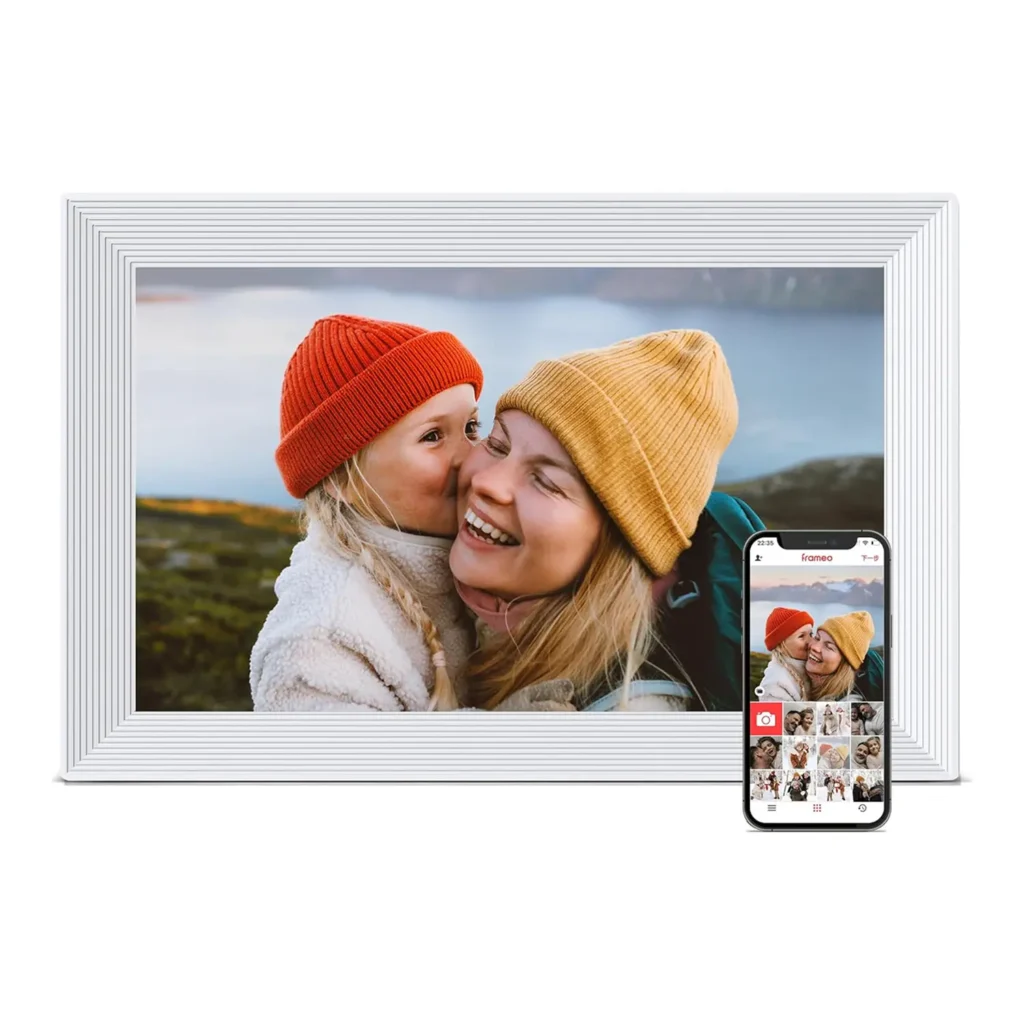 White digital photo frame showing a photo of a little girl in a red hat kissing her mother in a yellow hat, with mountains in the background and a smartphone in front for app control