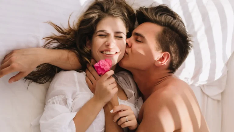 Happy couple in bed – woman in white shirt smiling with pink flower, man kissing her on the cheek