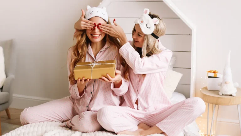 Two young girls in pink pajamas sitting on a white bed, one covering the other’s eyes while she holds a surprise gift, both wearing sleep masks, creating a cute and cozy bedroom moment perfect for friendship or gift-themed content