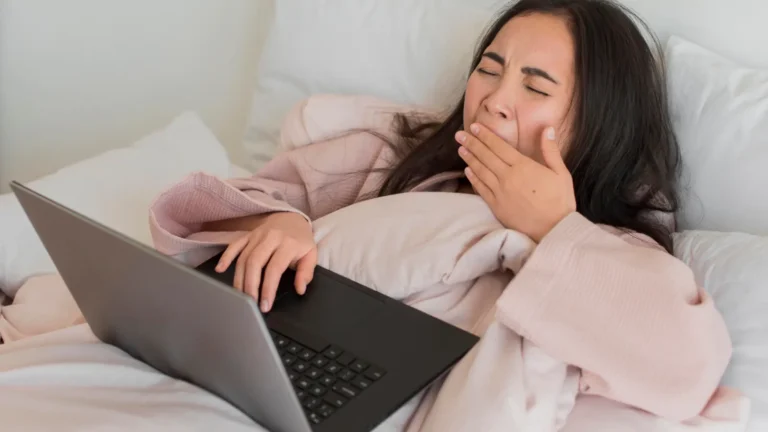 Young woman with black hair yawning while lying on a bed in a pink robe, laptop on her stomach, symbolizing lazy zodiac vibes.