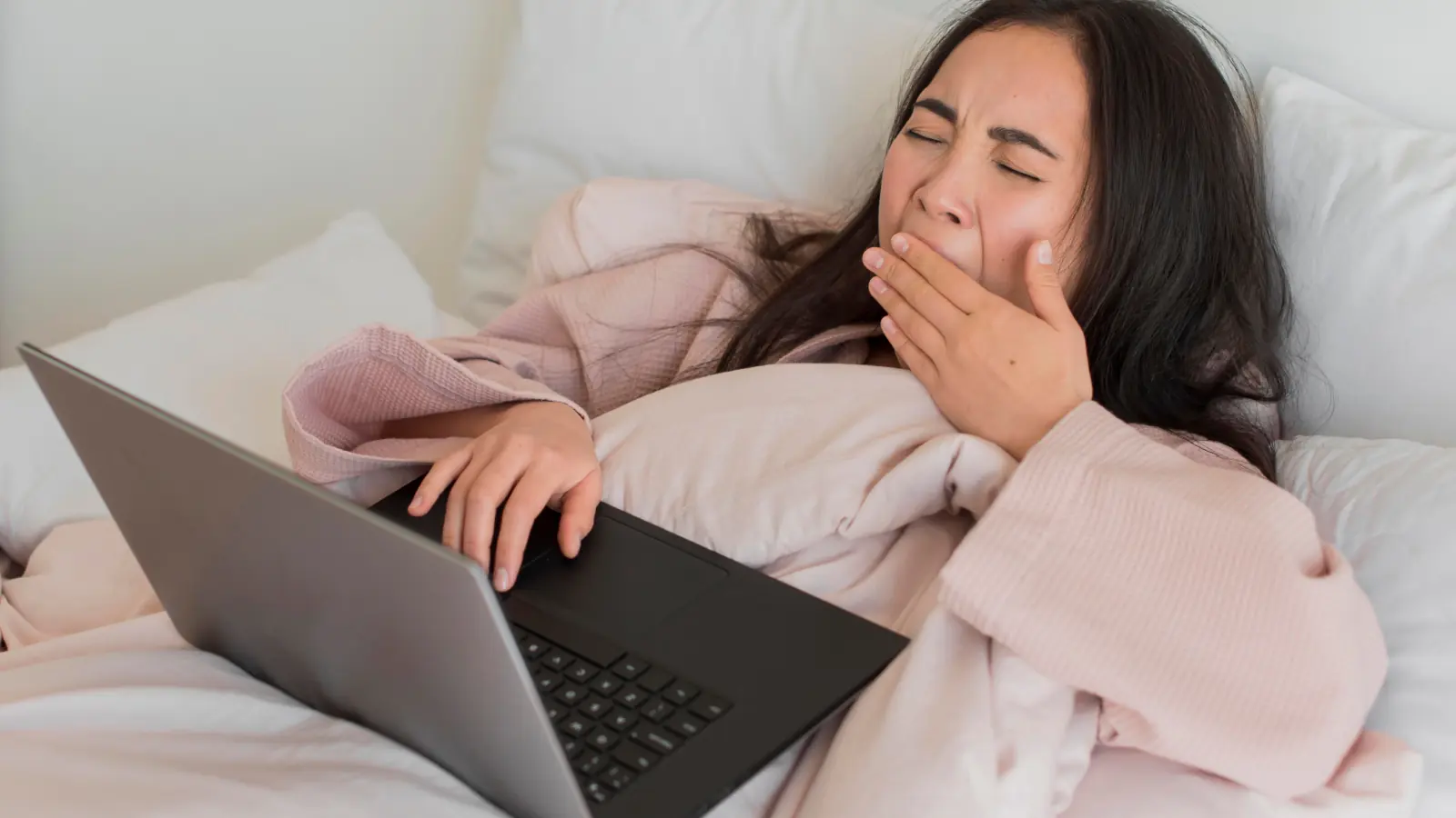 Young woman with black hair yawning while lying on a bed in a pink robe, laptop on her stomach, symbolizing lazy zodiac vibes.