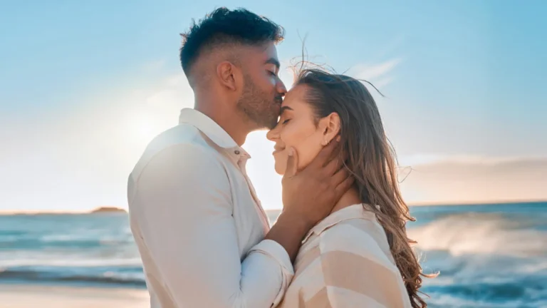 Man in a white shirt gently kissing woman with long light brown hair on the forehead by the sea under a blue sky