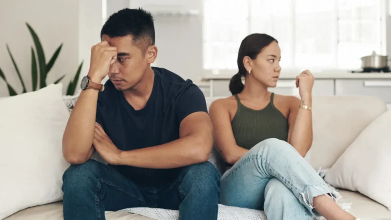 A young man and woman sitting on a beige sofa, with the man looking sad in the foreground, reflecting the pain of a breakup and emotional distance in their relationship