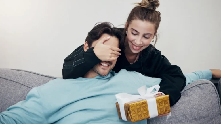Woman surprises her boyfriend on a gray couch, covering his eyes with one hand while offering a gold-wrapped gift with a white bow, both smiling in a playful, joyful moment.