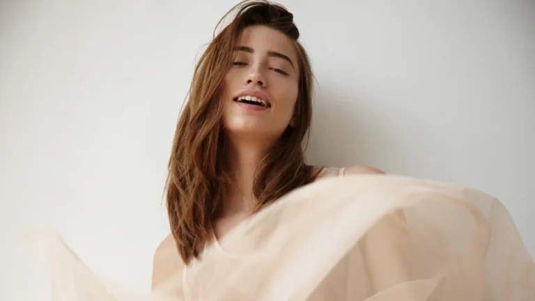 A stunning brunette woman with model-like features softly smiling, captured in an aesthetic photo with flowing beige fabric rising gracefully below her.
