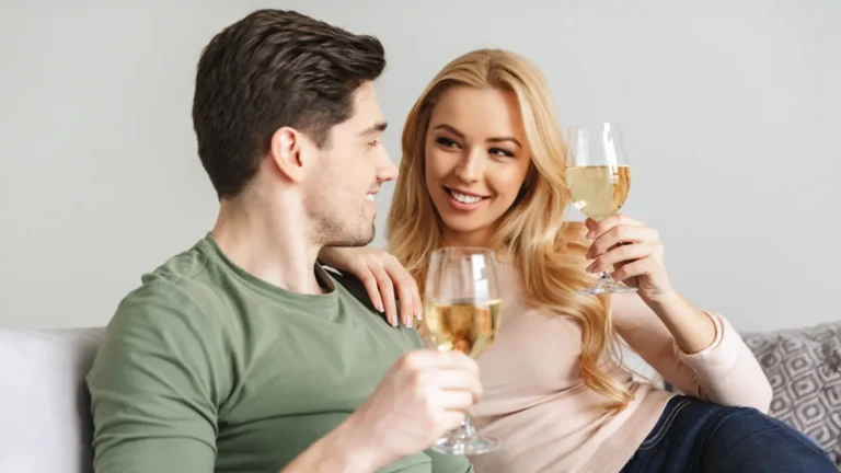 Smiling blonde woman in a pink sweater and dark blue jeans sits beside her brunette boyfriend in a pastel green sweater. They toast with champagne glasses while sitting close together on a gray couch against a light gray wall, looking at each other with a warm, flirty vibe.