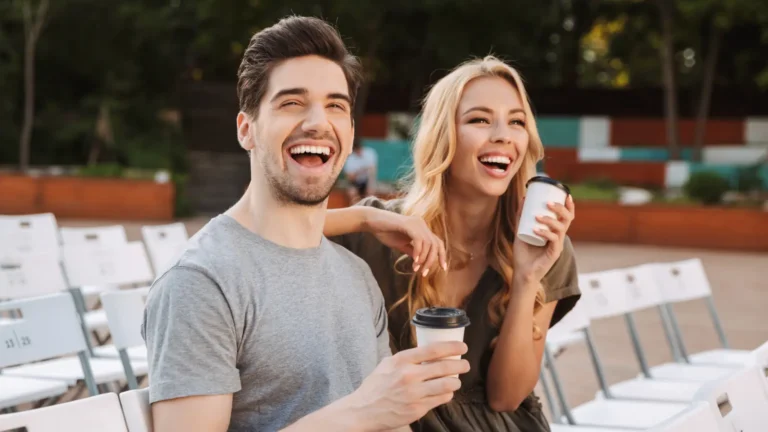 Happy brunette man and blonde woman sitting close together on white chairs outdoors, laughing and holding takeaway coffee cups surrounded by rows of white chairs.