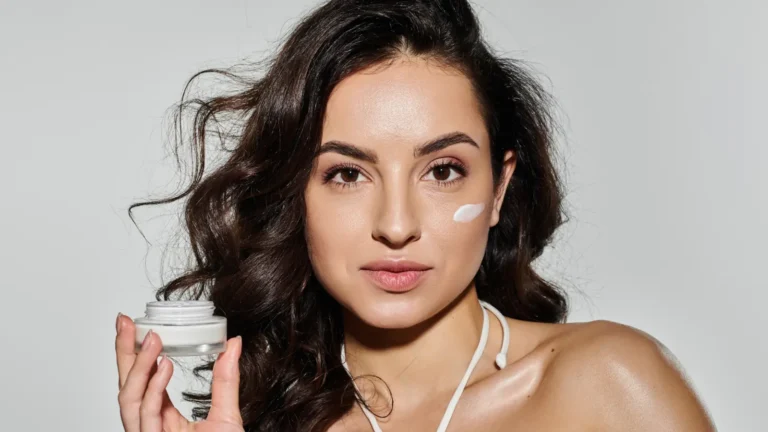 Young brunette woman with glowing skin holding a jar of face cream and applying it to her face, smiling and looking radiant